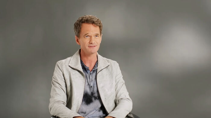 A person in a light jacket sits in a gray room, embodying celebrity style and demeanor.