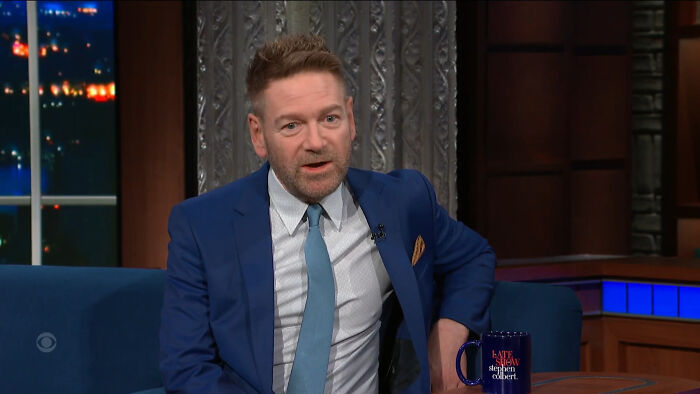 Man in a blue suit on a talk show set, discussing celebrities and petty reasons.