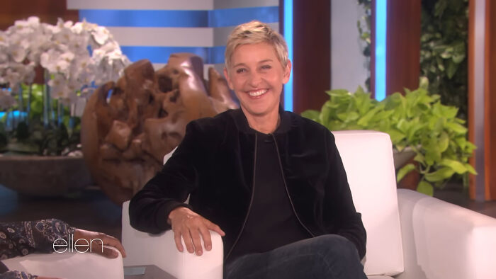 A person sits on a talk show set, smiling, with short blonde hair and wearing a black jacket.