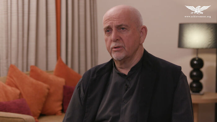 Bald man in black jacket discussing petty reasons in a cozy room with orange and red cushions and a table lamp.