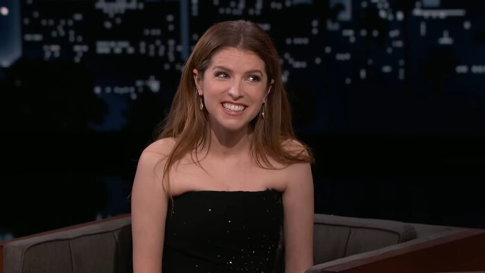 Celebrity smiling in a strapless dress during a talk show, cityscape backdrop.