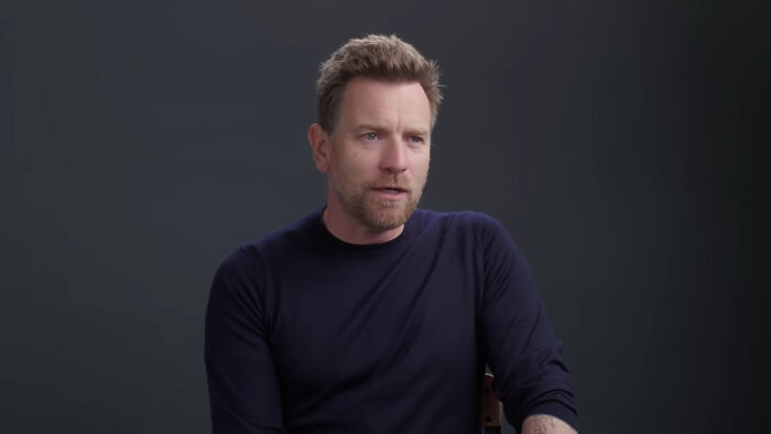 Man in a black sweater sitting against a dark background, the subject of celebrity discussion and controversies.