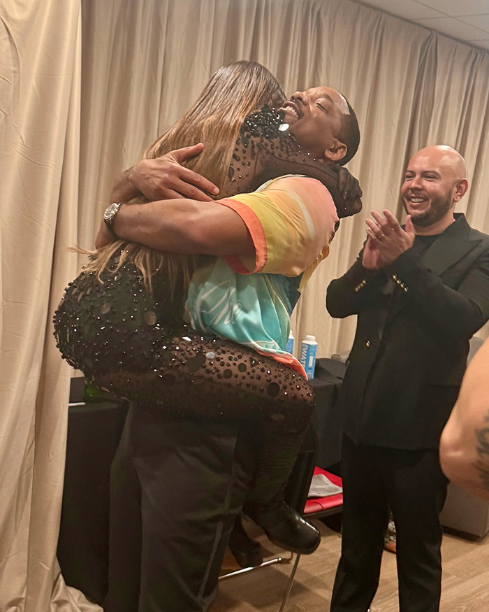 Man hugging a woman in a colorful shirt, with another man clapping, related to Will Smith controversy.