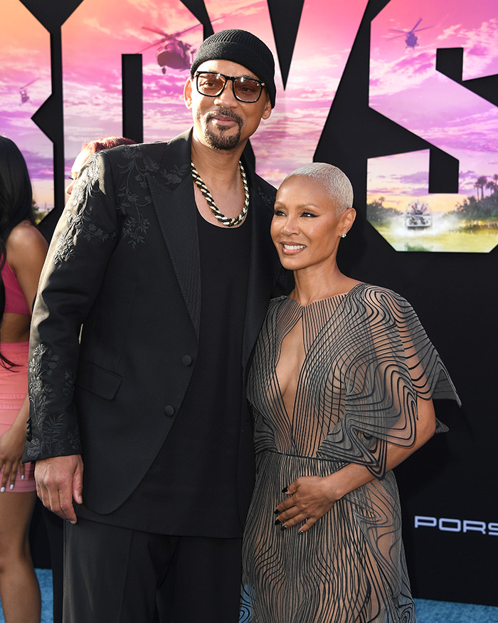 Man in a black suit and woman in a sheer dress posing together on a red carpet.