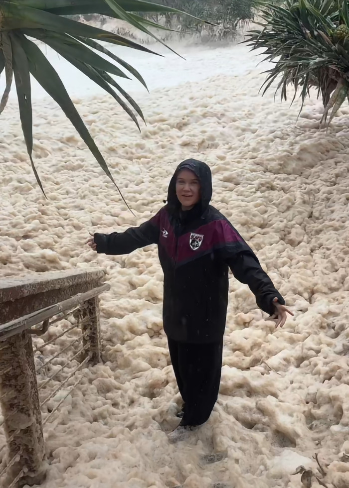 Woman in dark jacket standing amidst sewage-filled sea foam near a railing.