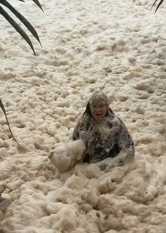 Woman performs stunt in sewage-filled sea foam, partially covered, amidst palm leaves, sparking outrage online.